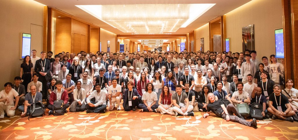 A large group of QTML participants gathered in rows in the hallway of the conference venue for a commemorative photo.
